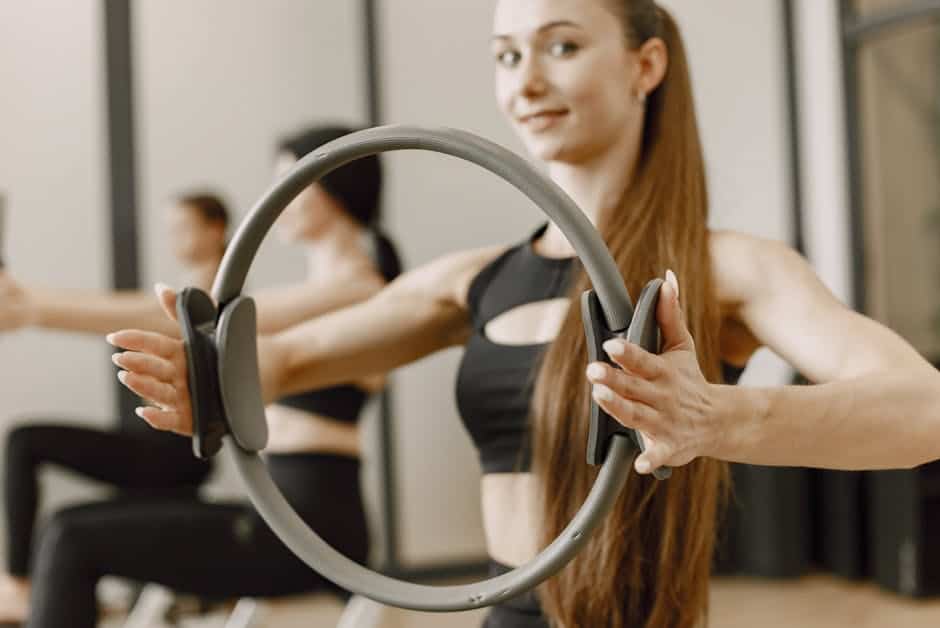 Woman in workout clothes holding a Pilates ring with both hands in a gym setting, with other people exercising in the background.